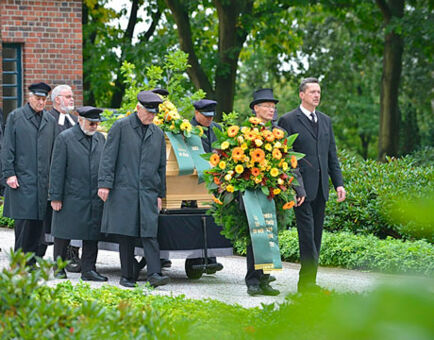 Das Bild zeigt einen Trauerzug mit einem Sarg. Vor dem Sarg wird ein großer schöner Kranz getragen.