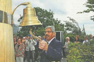 Eine Person läutet eine Glocke an einem Pfahl (Gedenkdalbe) vor zuschauenden Menschen.